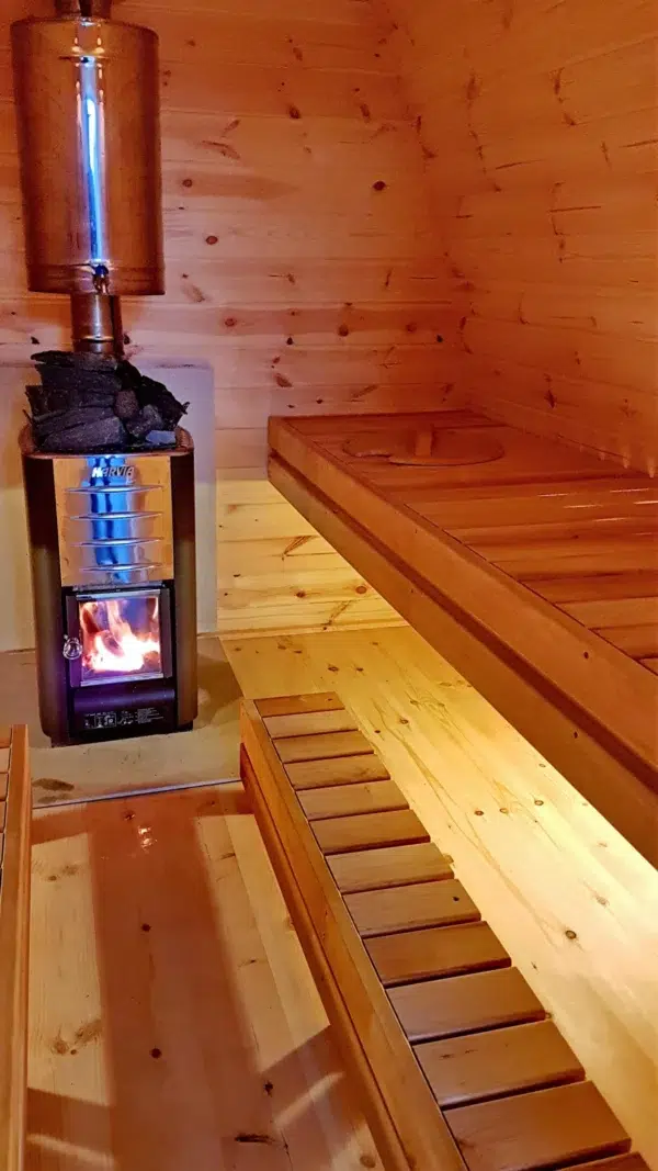 Outdoor pod sauna, lit wood-burning stove beside tiered benches